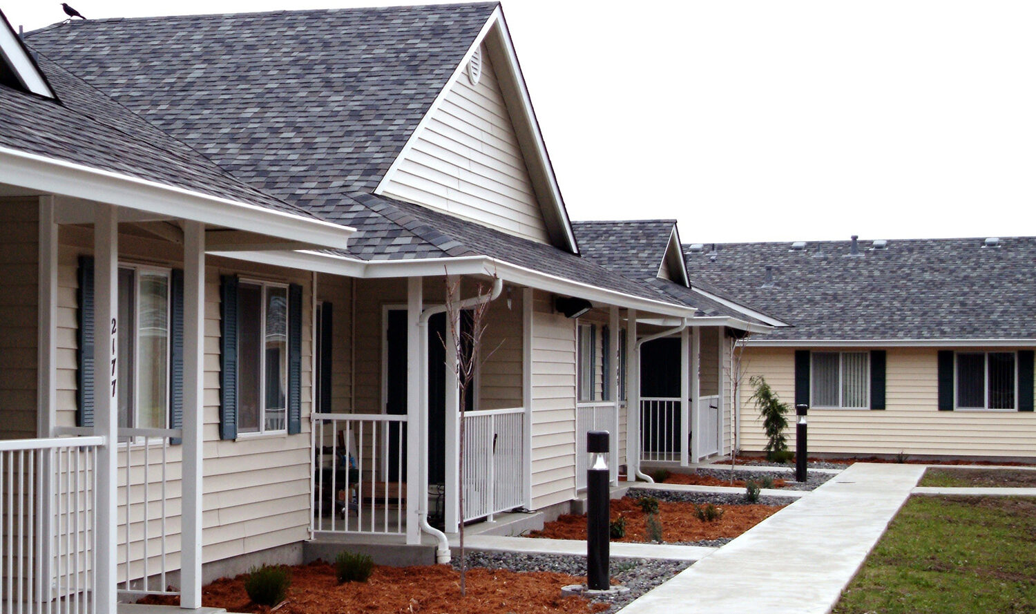 A row of single-story beige houses with gabled roofs and small front porches. Each house has an identical design, with landscaped front areas featuring mulch and small plants. A concrete pathway runs alongside the houses.