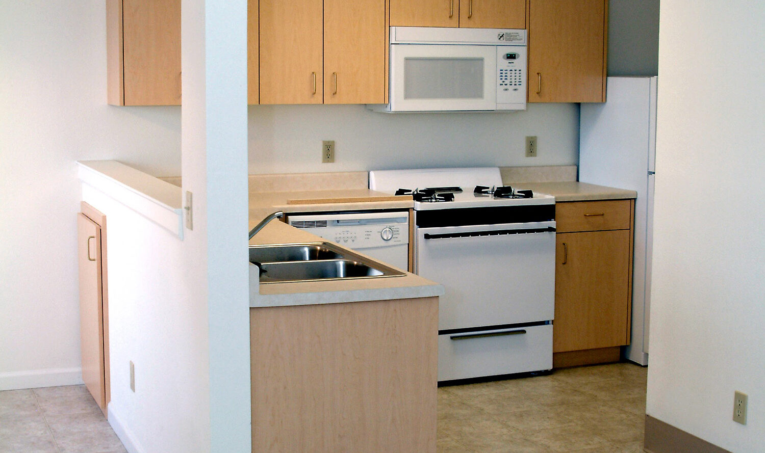 A small kitchen with light wood cabinets, a white stove, microwave, and refrigerator. The kitchen features a stainless steel sink and a small counter space. The room has tile flooring transitioning to carpet. Bright overhead lighting.