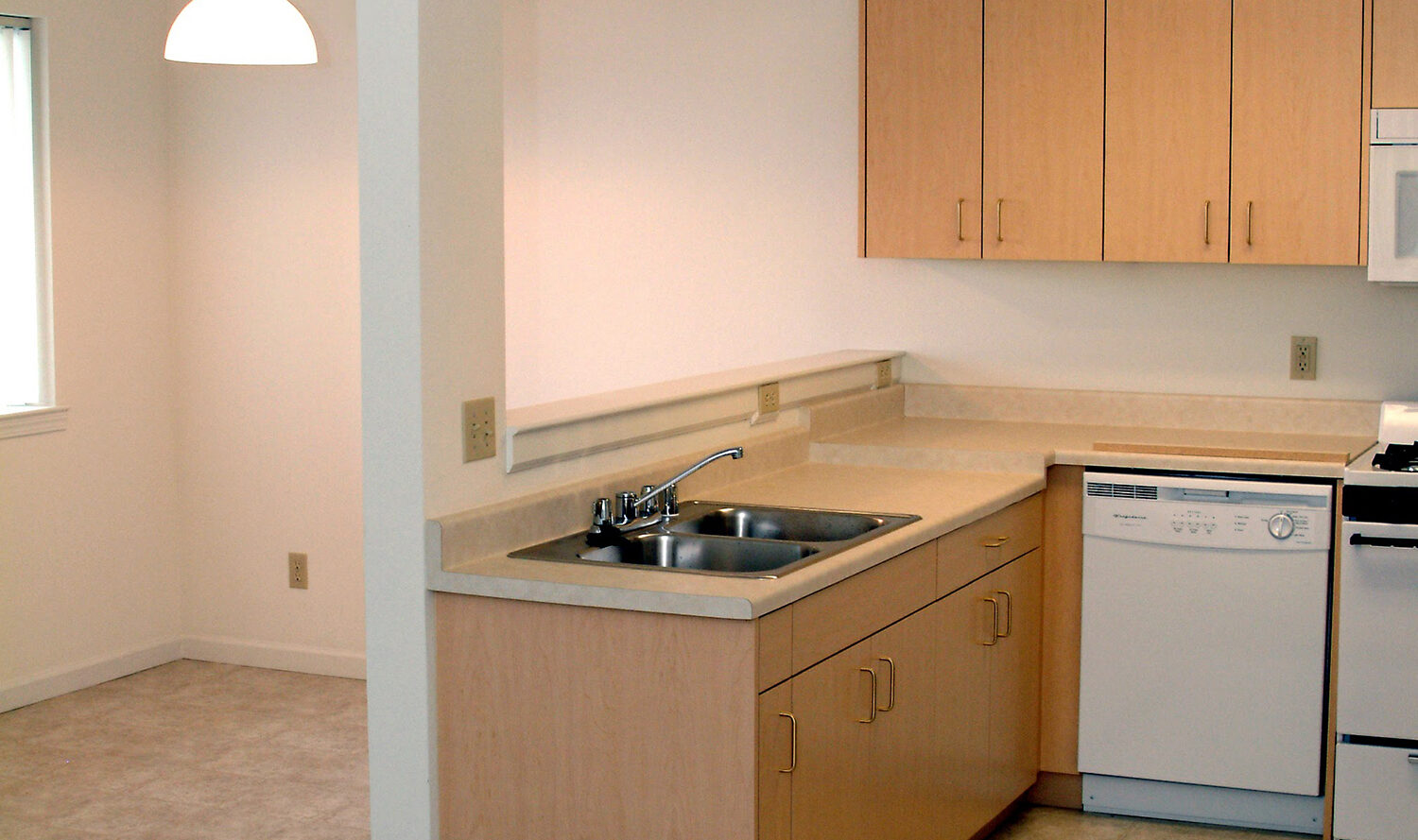 A modern, open-plan kitchen with light wood cabinets, a sink, dishwasher, and oven. The kitchen features tiled flooring, a carpeted adjoining area, and a hanging ceiling light in the dining space. The overall color scheme is neutral.