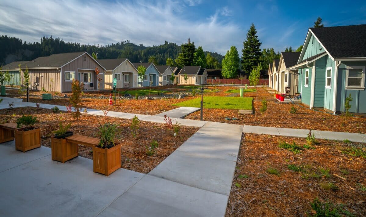 A peaceful suburban neighborhood with several small, colorful houses surrounded by landscaped gardens. A concrete pathway runs through the area, with wooden planters placed along it. The background features a forested hill under a blue sky.