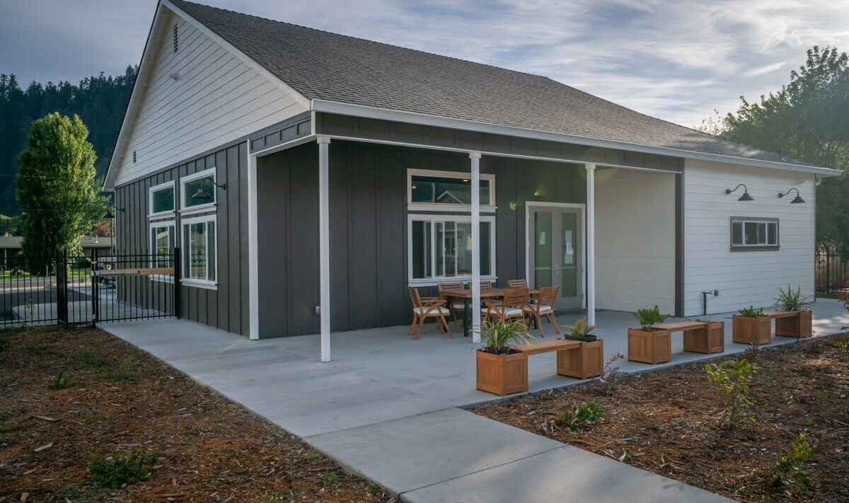 A modern single-story building with dark and light siding sits under a partly cloudy sky. Theres a small patio with a table and chairs, surrounded by planters. Paths and sparse landscaping surround the building.