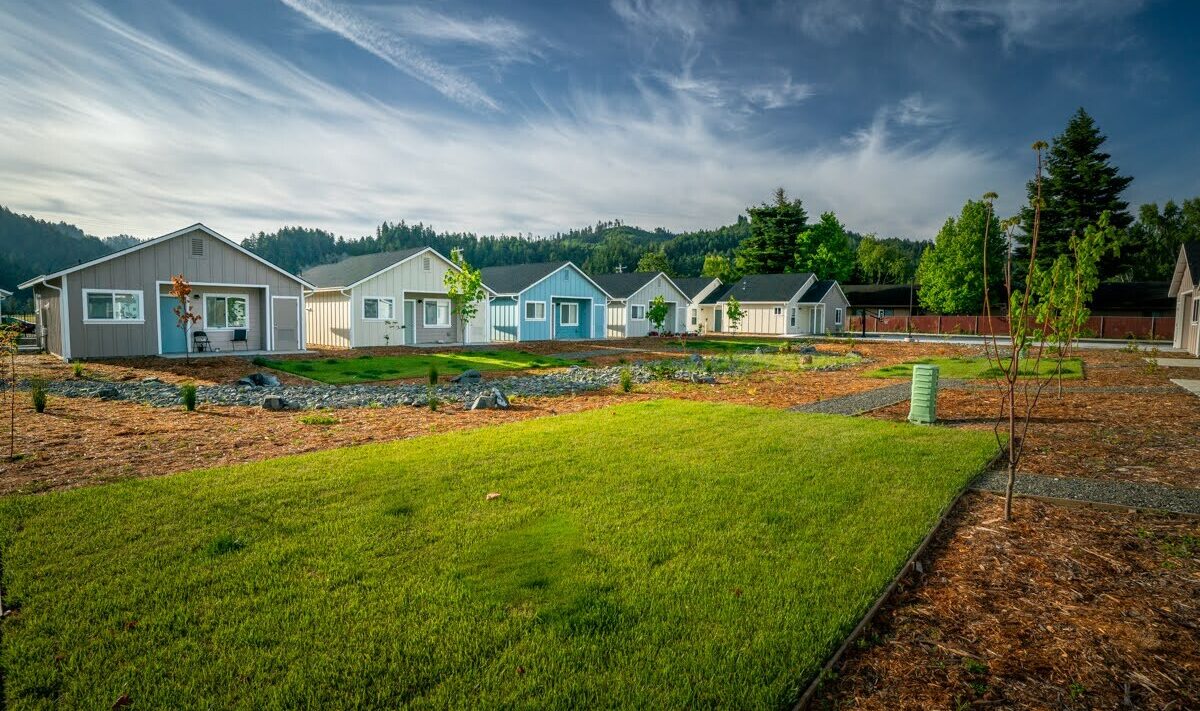 A row of small, colorful houses lines a lush green lawn under a partly cloudy sky. Trees and a garden area are visible in the background, creating a serene neighborhood scene.