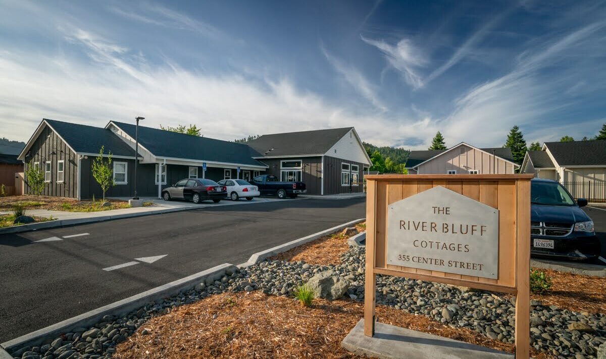 A row of modern cottages under a clear blue sky, with several cars parked in front. A sign in the foreground reads The River Bluff Cottages, 355 Center Street. Surrounded by greenery and a neatly paved driveway.