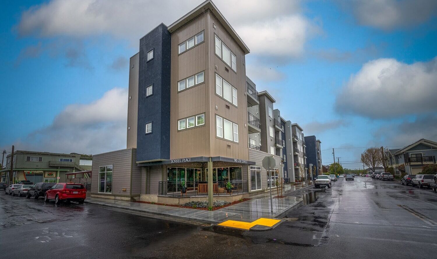 A modern multi-story building on a rainy day with a cloudy sky. The building has a mix of beige and dark blue panels, large windows, and a small storefront at the corner. Several parked cars line the wet street.