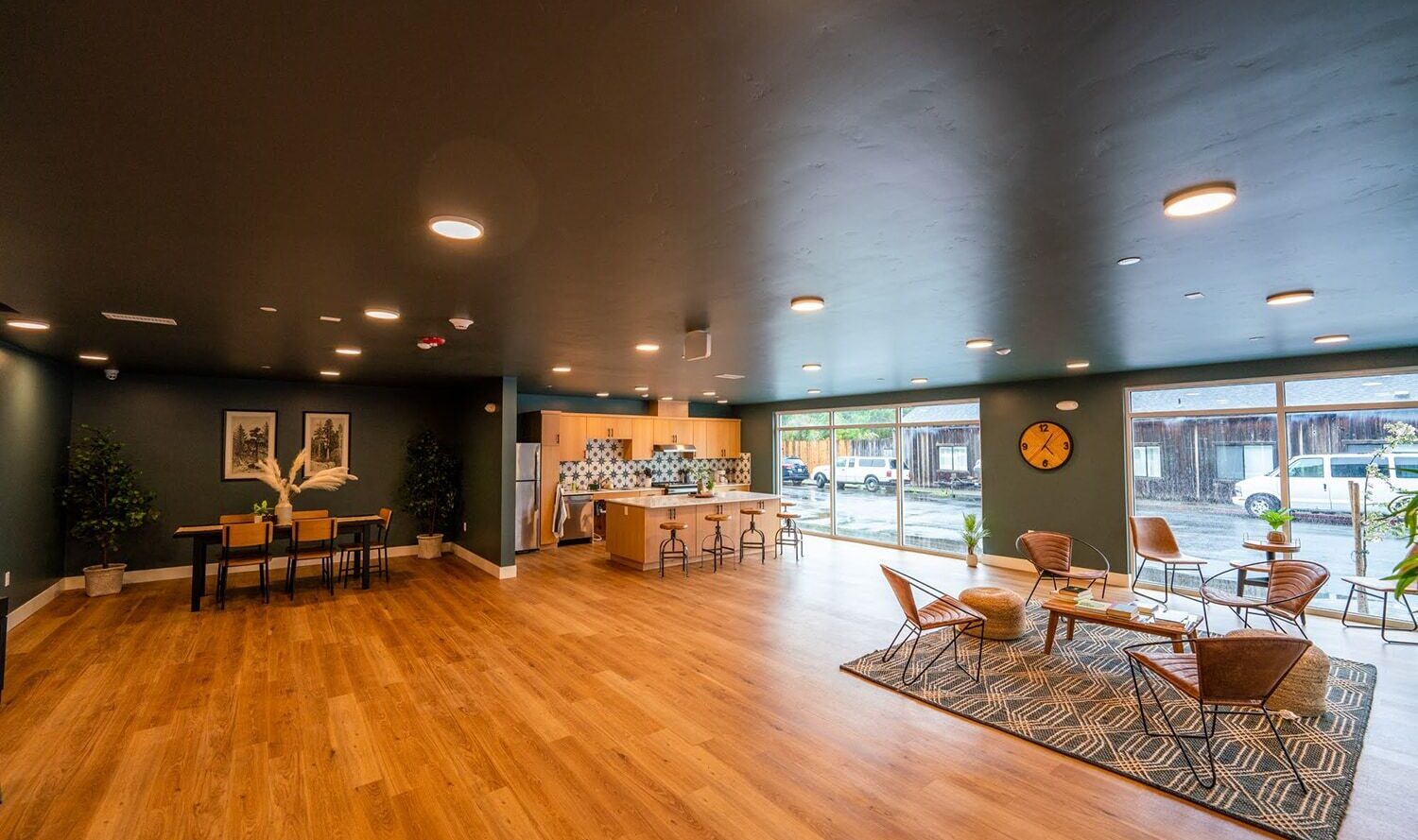 A spacious, modern room with wooden flooring, featuring a sitting area with wicker chairs and a rug. A kitchen is visible in the back, along with a high table and stools. Large windows let in natural light, and there are plants placed throughout.