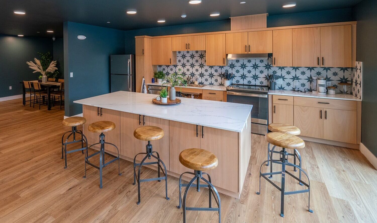 A modern kitchen with light wood cabinets, a large island with stools, and patterned tile backsplash. Stainless steel appliances and wooden flooring complete the look. A dining area with a table and chairs is visible in the background.