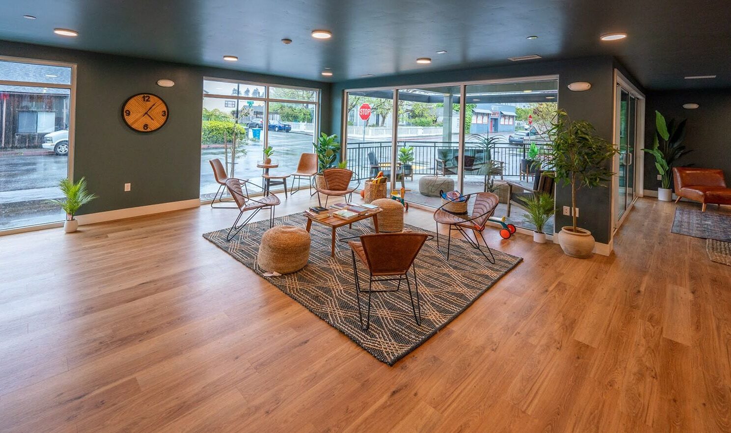 A modern, open lounge area with wooden flooring, featuring a circular clock on a dark wall. It has a seating arrangement with brown chairs, a rug, potted plants, and large windows offering a view of the outside. Bright and inviting atmosphere.