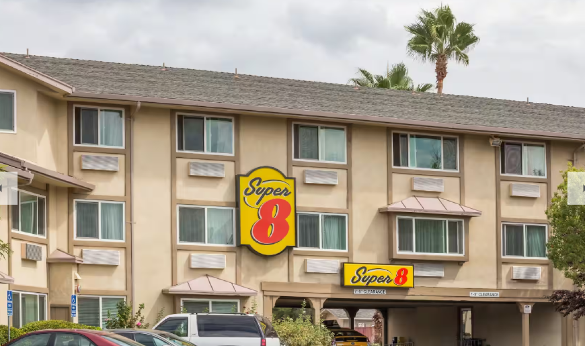 Front view of a beige three-story hotel with a Super 8 sign. The building has palm trees nearby and multiple windows, each with an awning. The sky is cloudy.