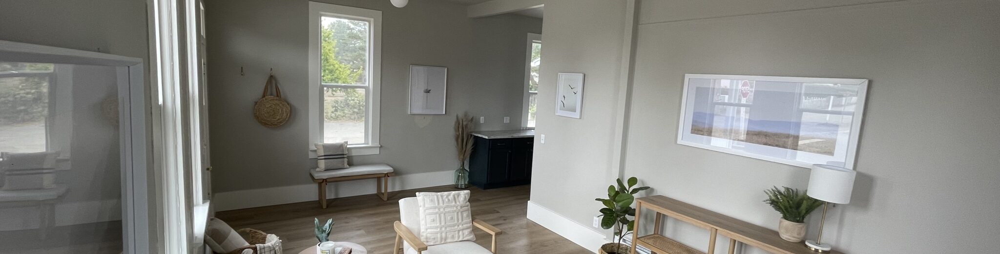 A modern living room with neutral tones, featuring a gray sofa, wooden chairs with cushions, a white ceiling fan, and a light rug. There are framed artworks on the walls, a potted plant on a console table, and large windows providing natural light.