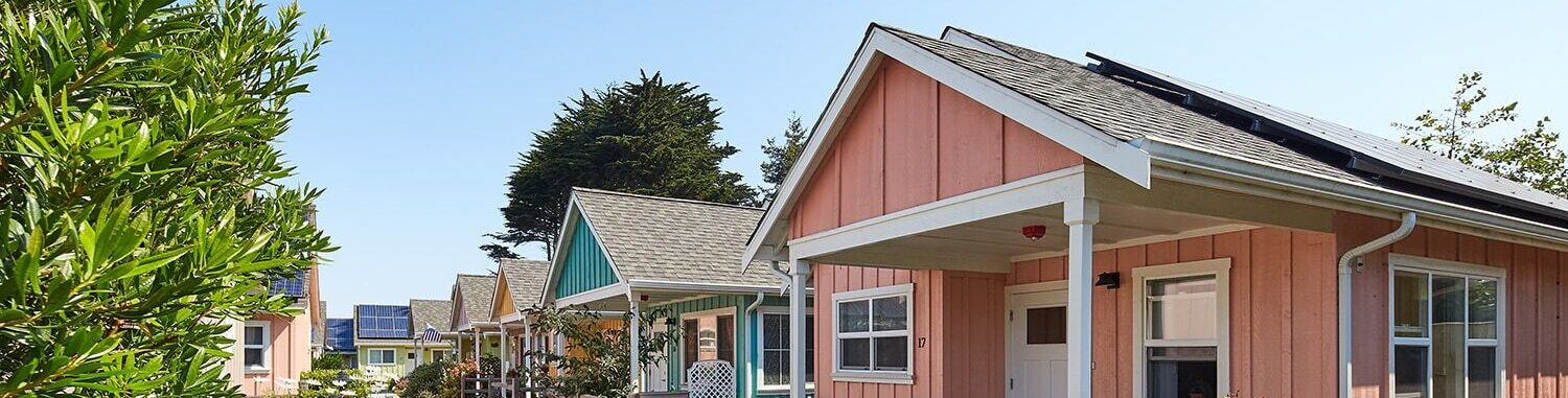 A row of small, colorful houses with well-maintained gardens line a sunny street. Solar panels are visible on the roofs, and a clear blue sky is in the background, suggesting an environmentally friendly neighborhood.