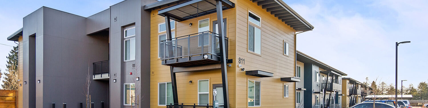 Modern apartment complex with two stories, featuring yellow and gray exterior panels. The building has balconies and large windows. A parking area with a few cars is in the foreground, and the sky is clear and blue.