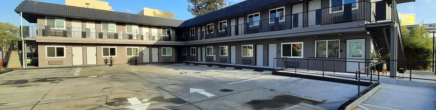 A two-story motel with an empty parking lot under a clear blue sky. The building has a dark roof, beige walls, and a few scattered trees in the background. A wheelchair ramp is visible on the right side.