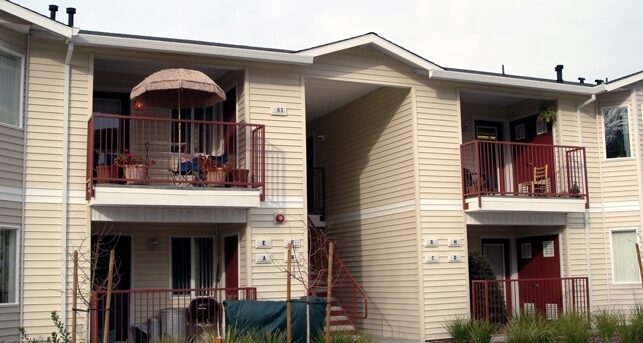 A two-story apartment building with beige siding and red railings. The upper floor has balconies with chairs and a parasol. The entrance is centered, leading to a staircase. Green plants are in front of the building.