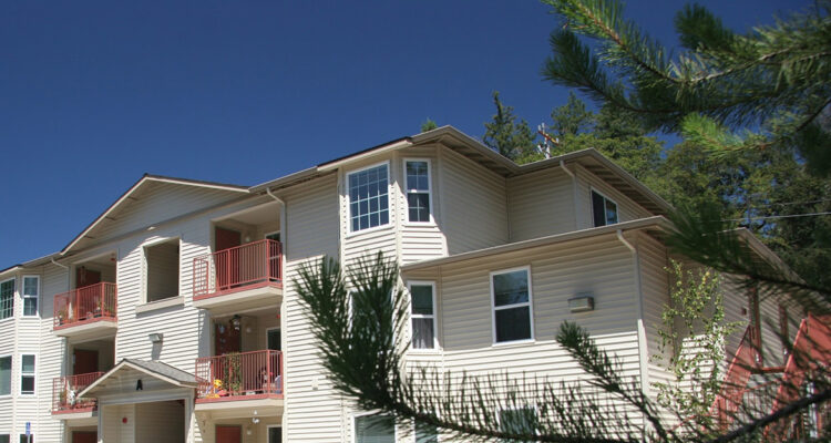Three-story residential building with beige siding and red balconies, surrounded by pine trees under a clear blue sky.