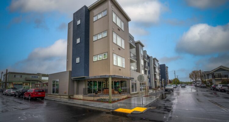 A modern, multi-story apartment building stands at a street corner on a rainy day. The exterior features a mix of gray and beige panels. A few parked cars are visible along the wet street, and the sky is cloudy.