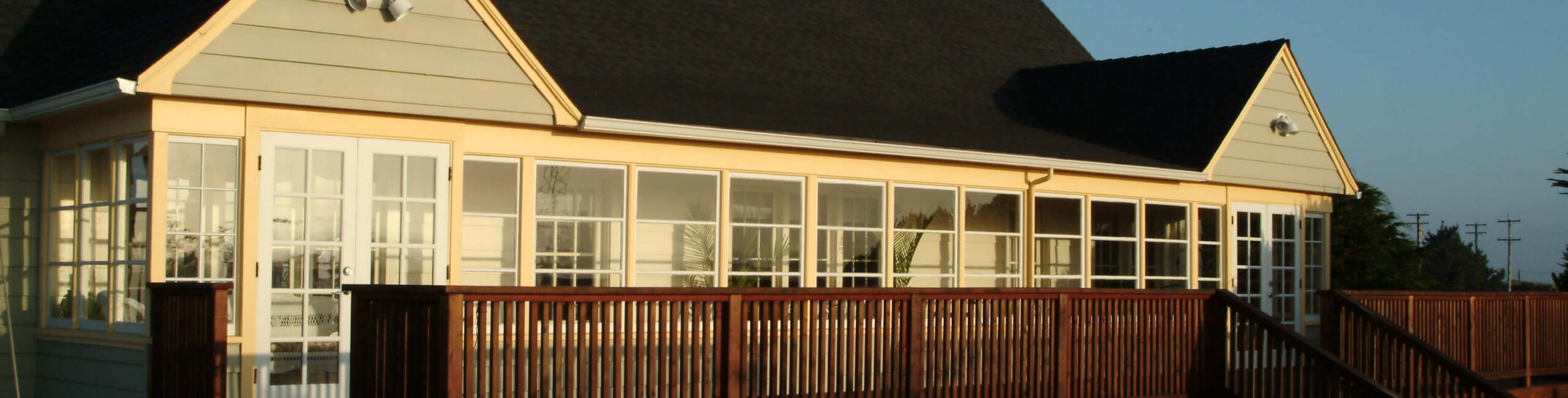 A single-story house with yellow siding and a dark shingled roof features a wooden deck and multiple windows. The house is illuminated by sunlight, and the sky is clear and blue.